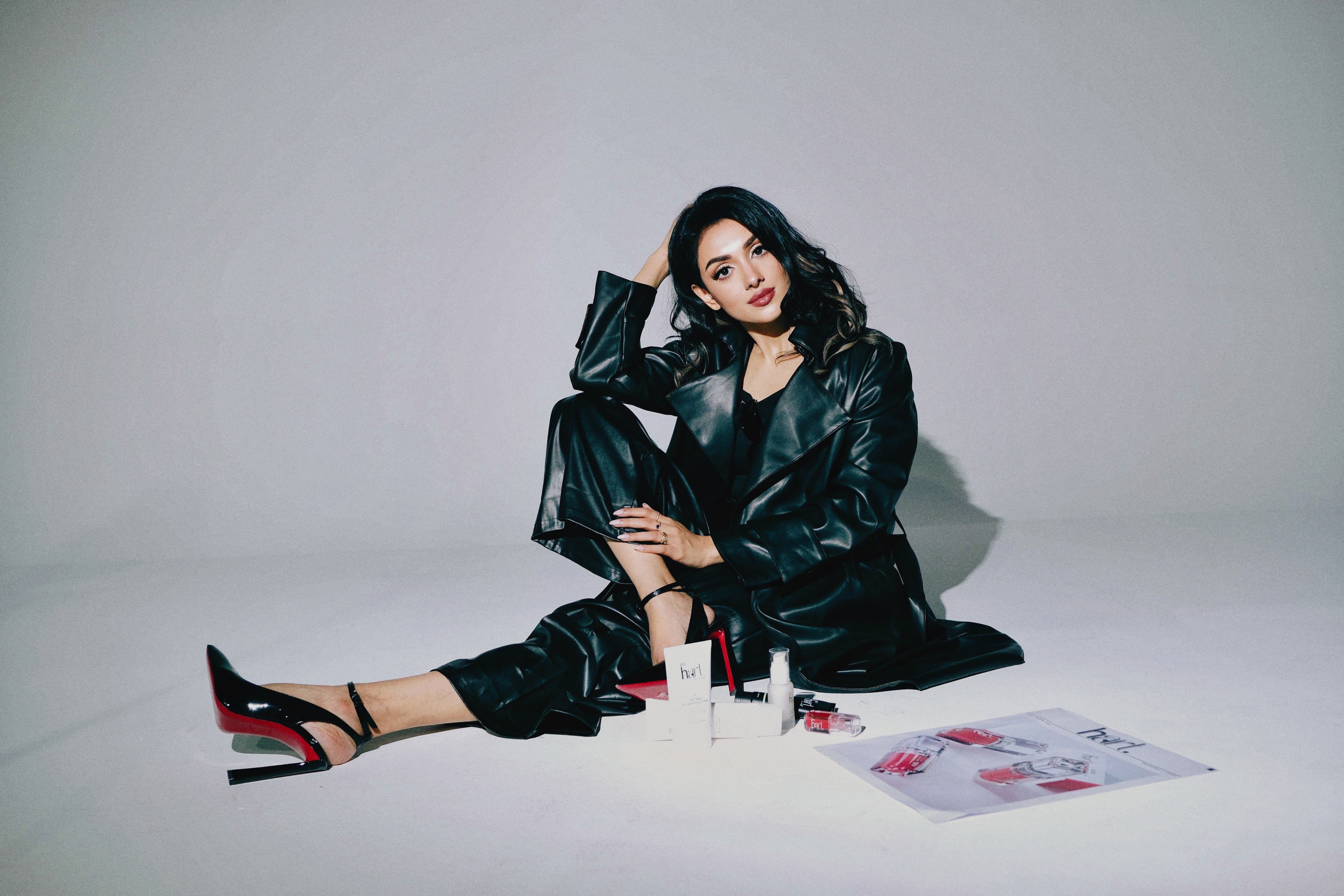 Woman in black leather outfit sitting on a white floor with makeup products around her.