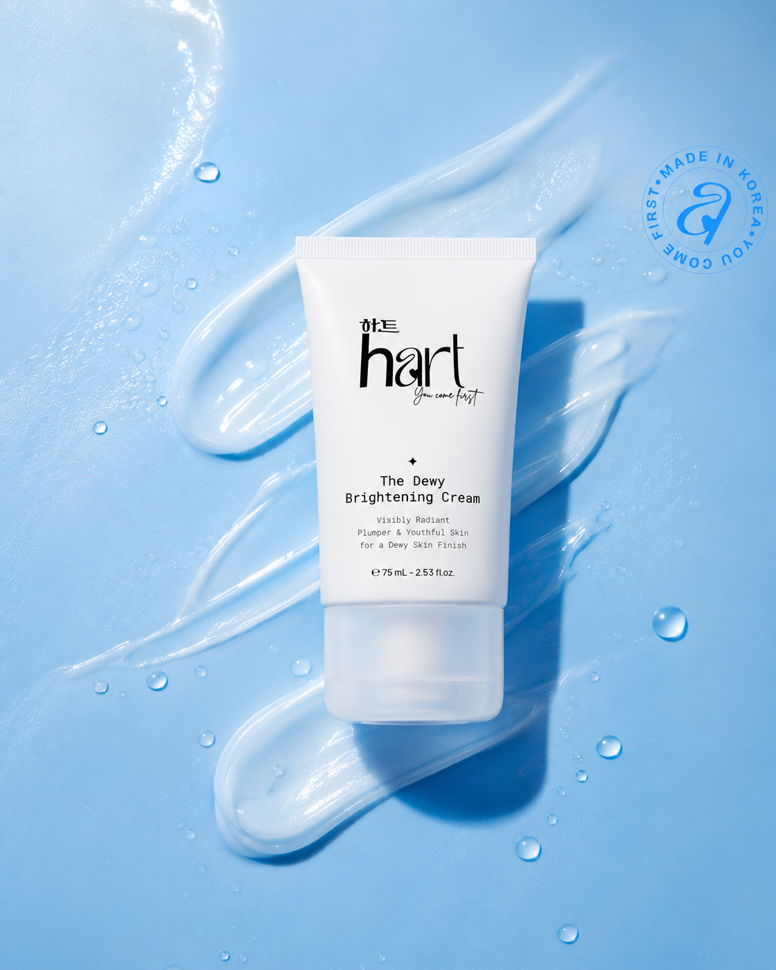 A white squeeze tube of 'the hart' The Dewy Brightening Cream centered on a light blue background with decorative cream swatches and water droplets. A circular 'First Made in Ontario' logo is visible in the top right corner.